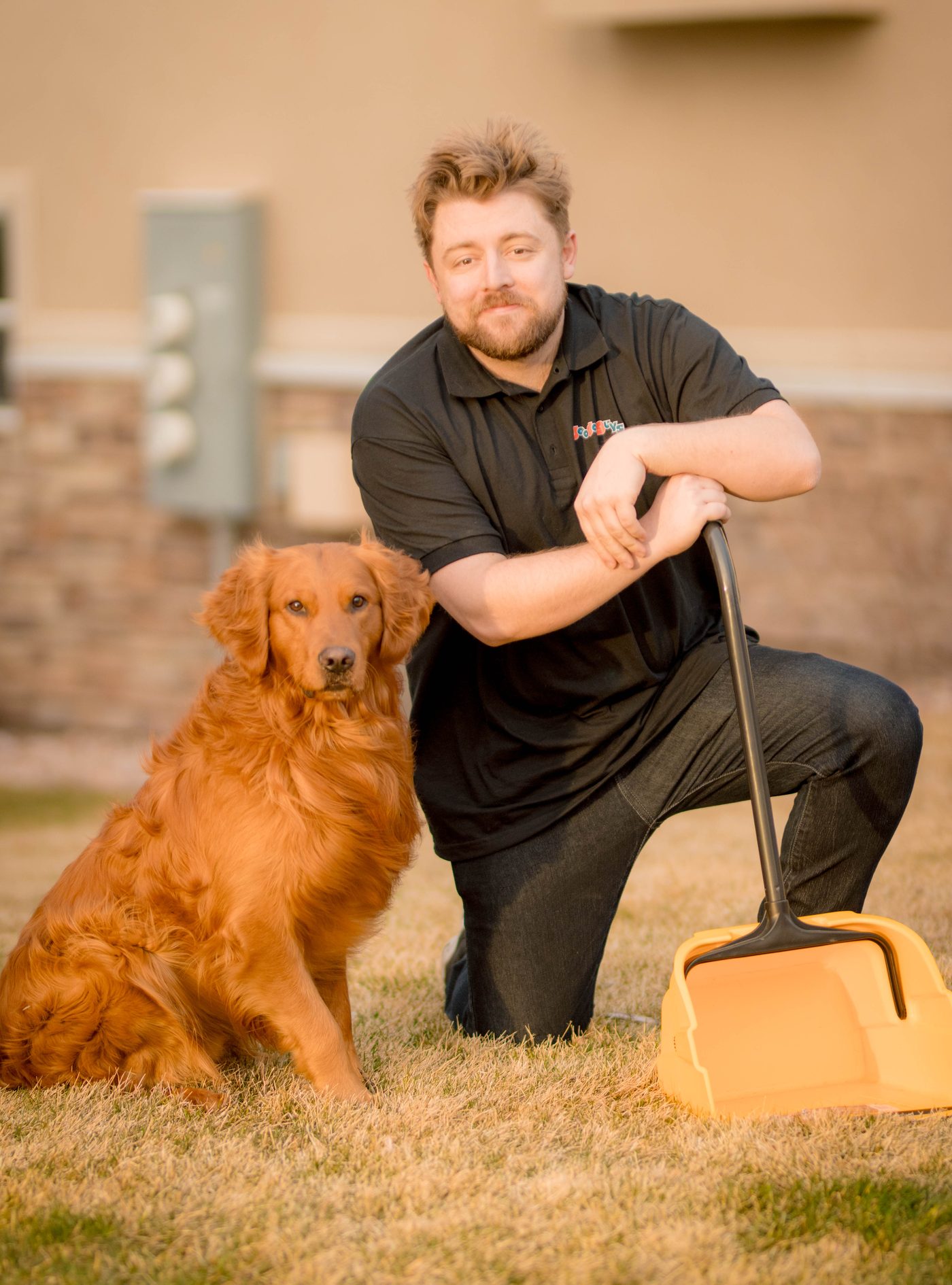 A Doodoo Guys technician kneeling with a golden retriever and scoop in a clean yard